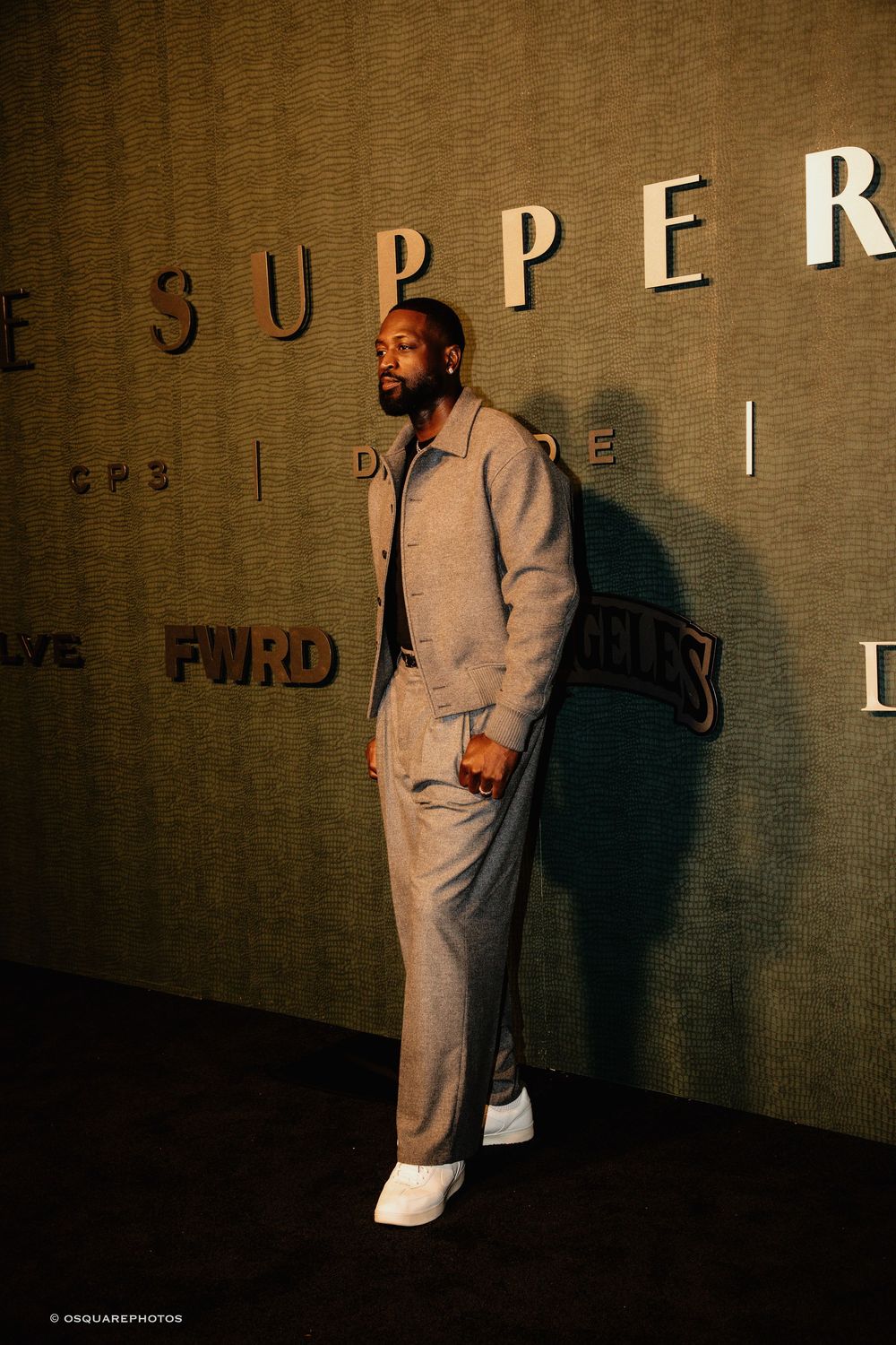 A person in a beige suit and white sneakers stands against a wall with 'SUPPER' text in dramatic lighting.