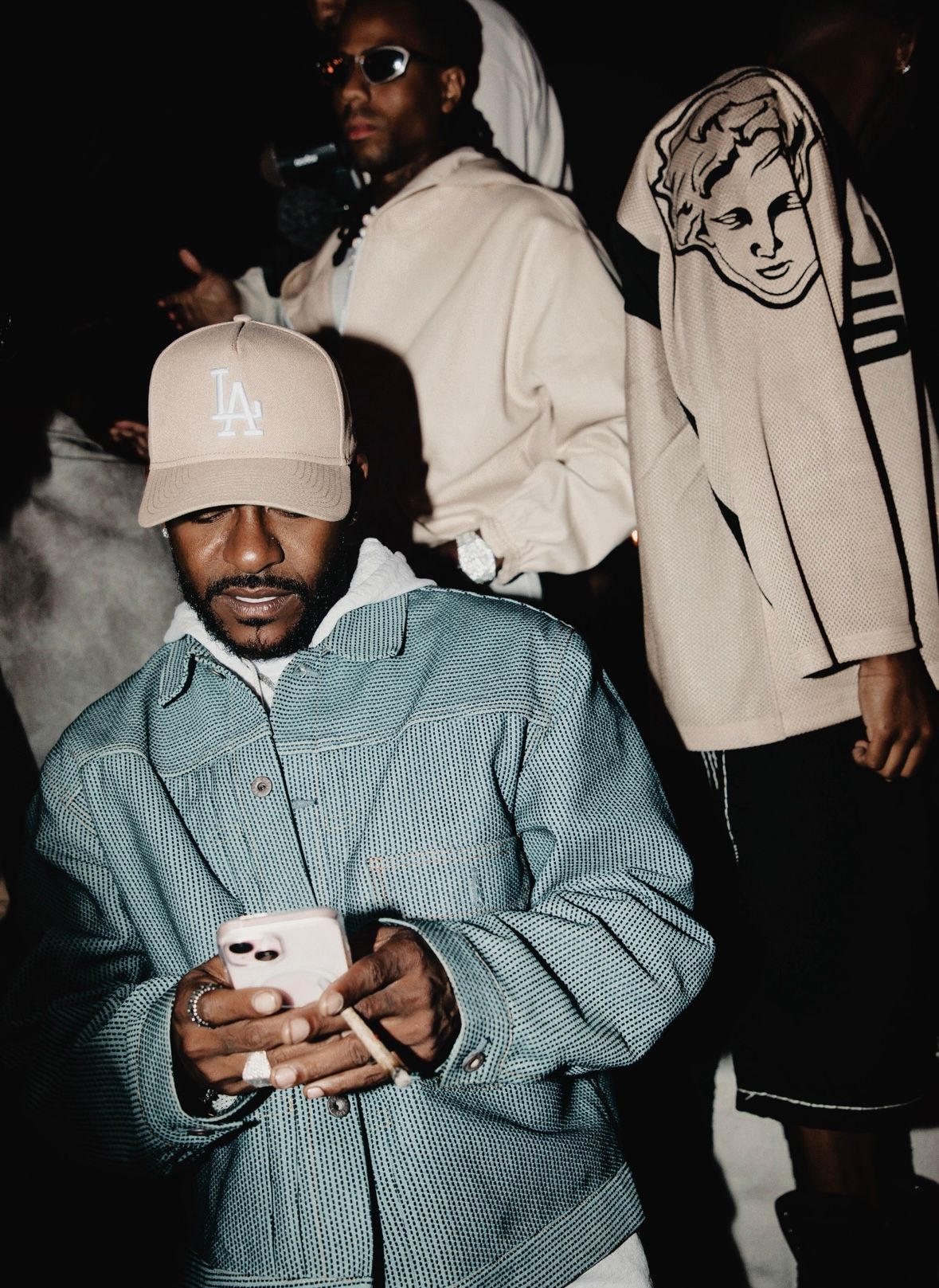 Two people in casual streetwear stand indoors, one wearing a blue corduroy jacket and LA cap checking their phone.