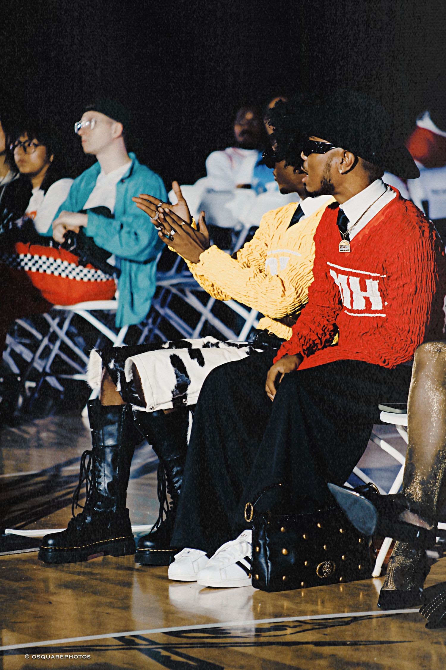 Row of people sitting on a gym floor wearing sneakers and casual clothing at an indoor event.