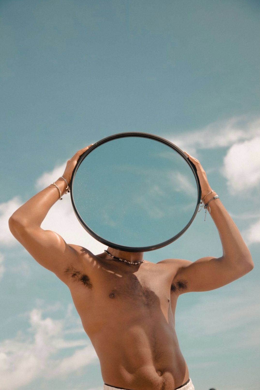Person holding a circular mirror against blue sky, creating artistic reflection composition.