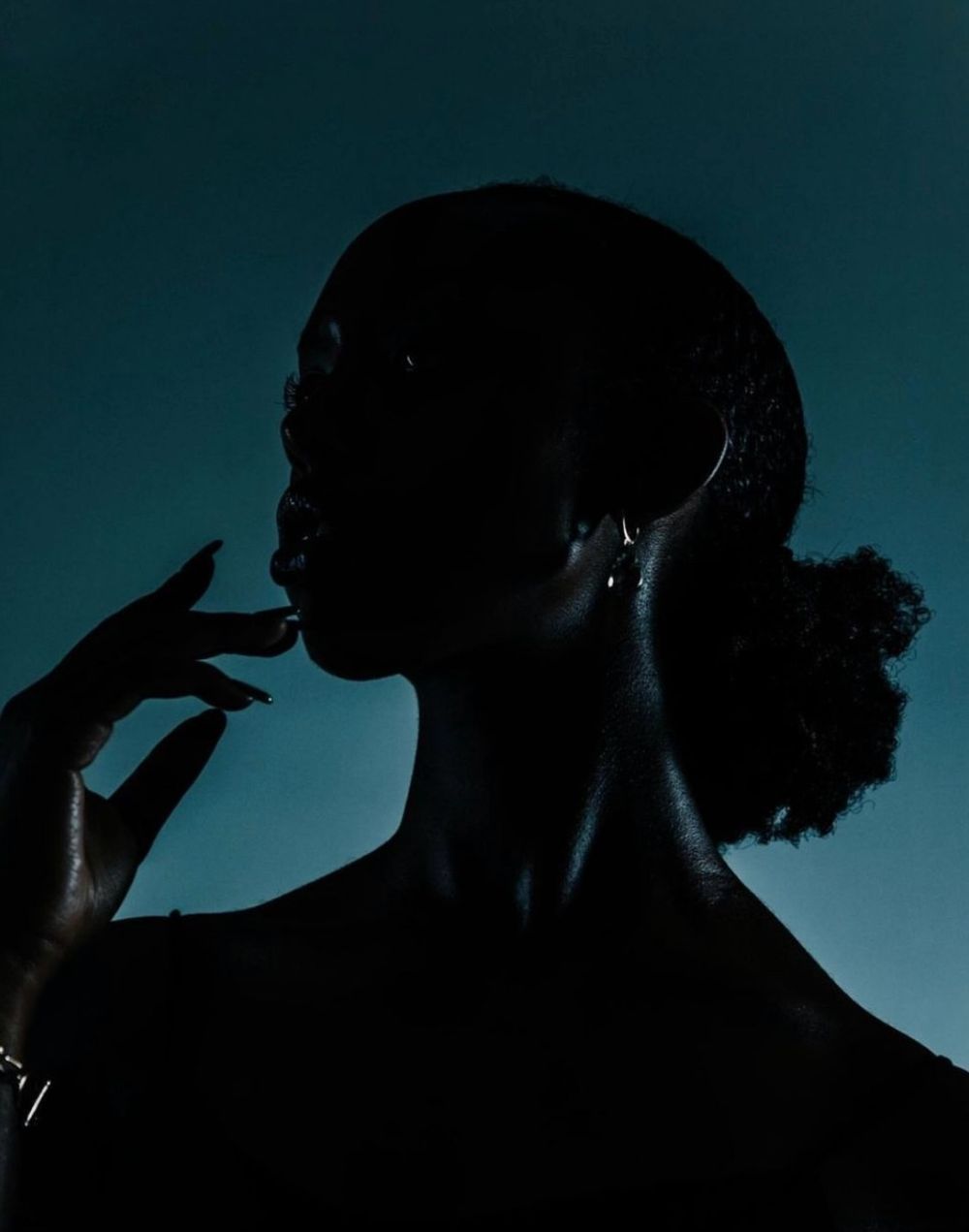 Silhouetted profile against a teal background shows a figure with natural hair gesturing near their face in dramatic lighting.
