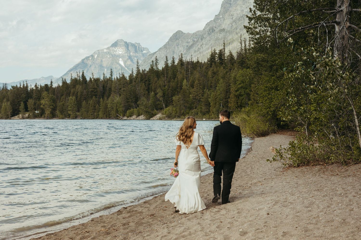 Maternity photo of couple standing on rocky shoreline with mountains and lake in background.