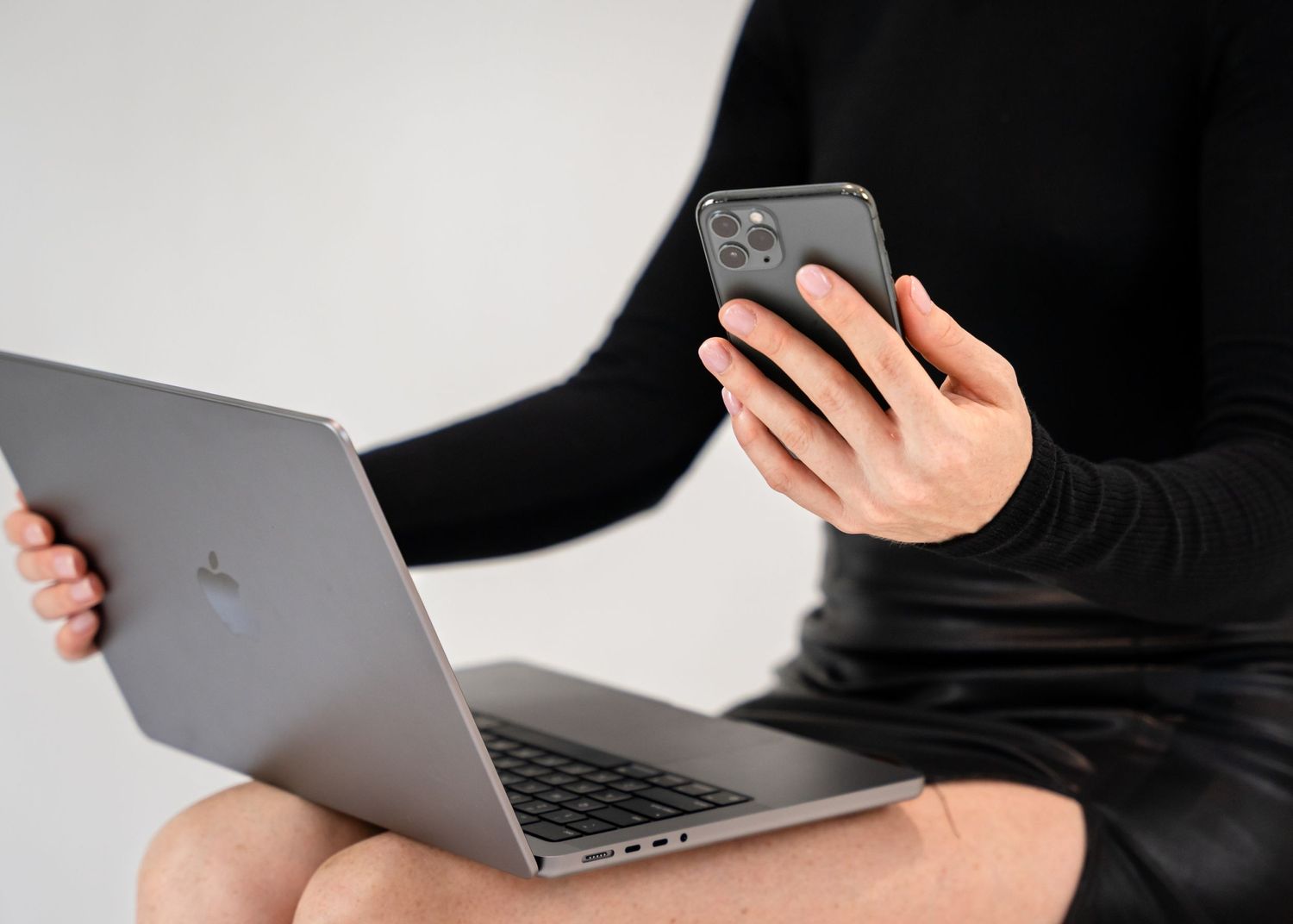 Laptop sitting on lap, holding iPhone in hand wearing black long sleeved shirt and skirt