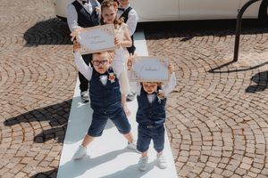 Due bambini con gilet e papillon portano cartelli nuziali lungo la navata all’aperto.