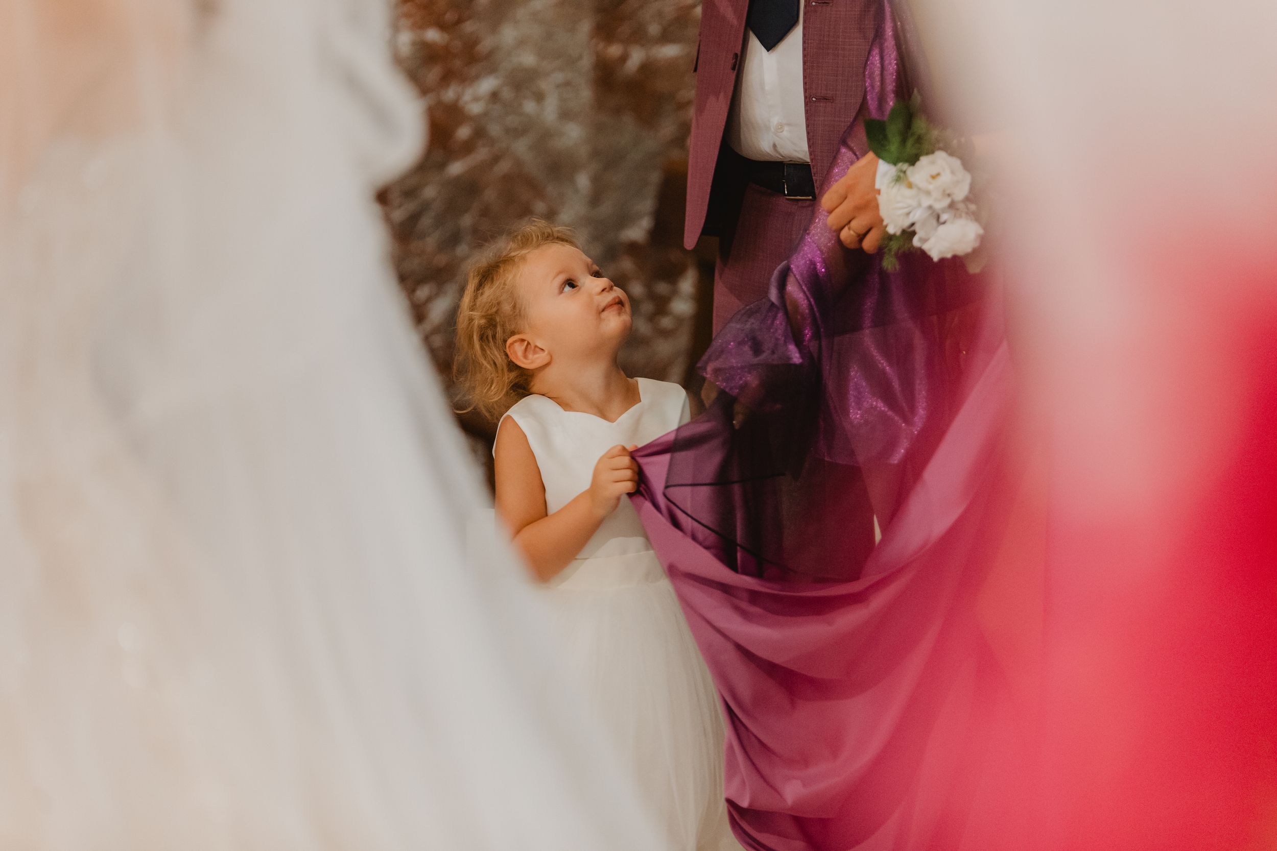 Una bambina in abito bianco guarda in alto stringendo il tessuto di un vestito viola.