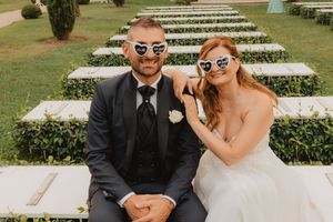 Gli sposi sorridono con occhiali a forma di cuore su gradini di pietra in giardino.