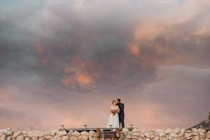 Salvatore e Mariarita in piedi su un muretto, sotto un cielo di nuvole rosa e viola al tramonto.