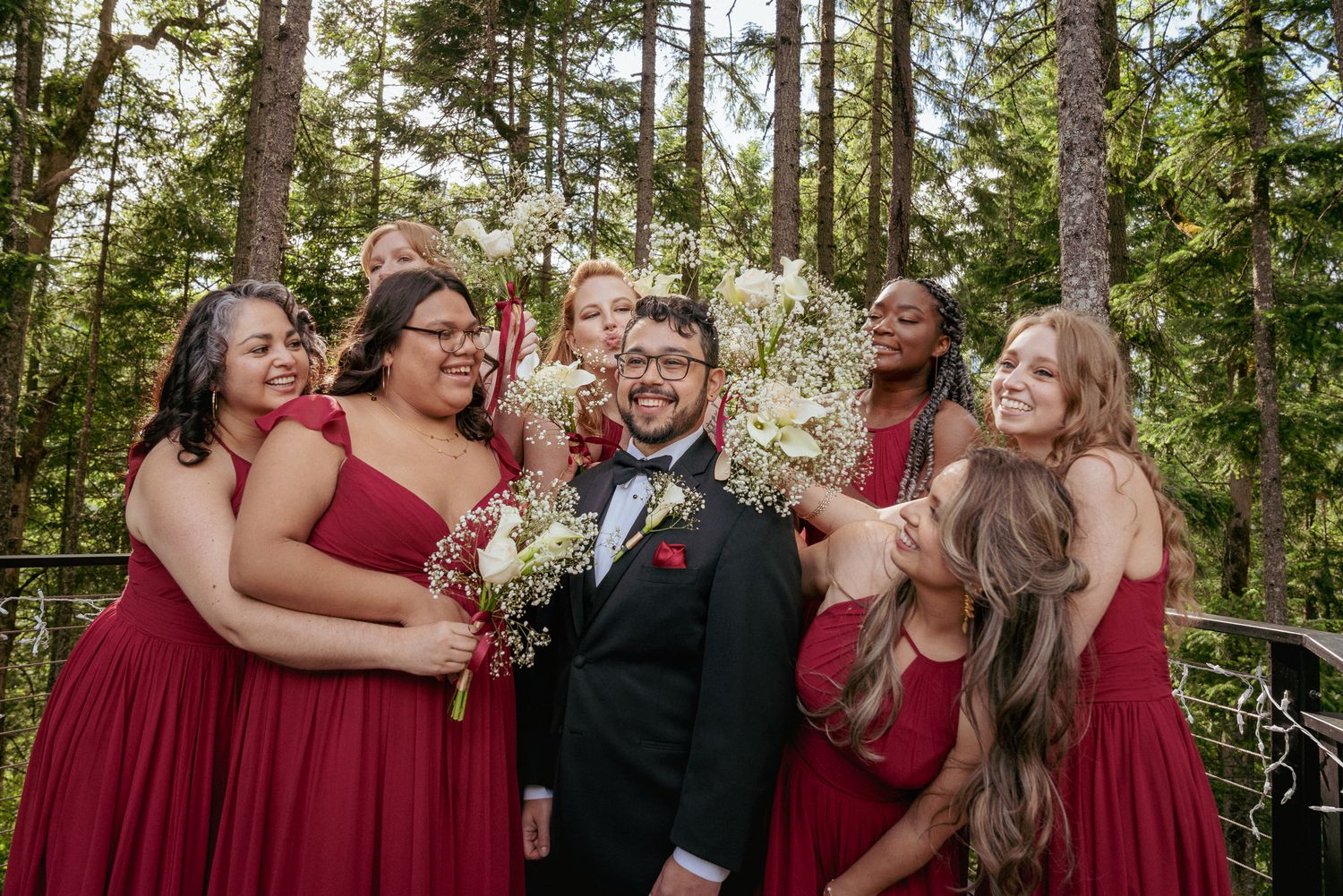 Groom surrounded by the bridesmaids that are putting bouquets around grooms face and laughing in PNW forest setting during outdoor wedding.