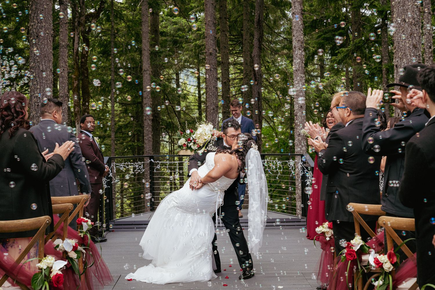 Interracial wedding ceremony in the forest as the newlyweds share a kiss while guests cheer and toss white confetti in celebration.