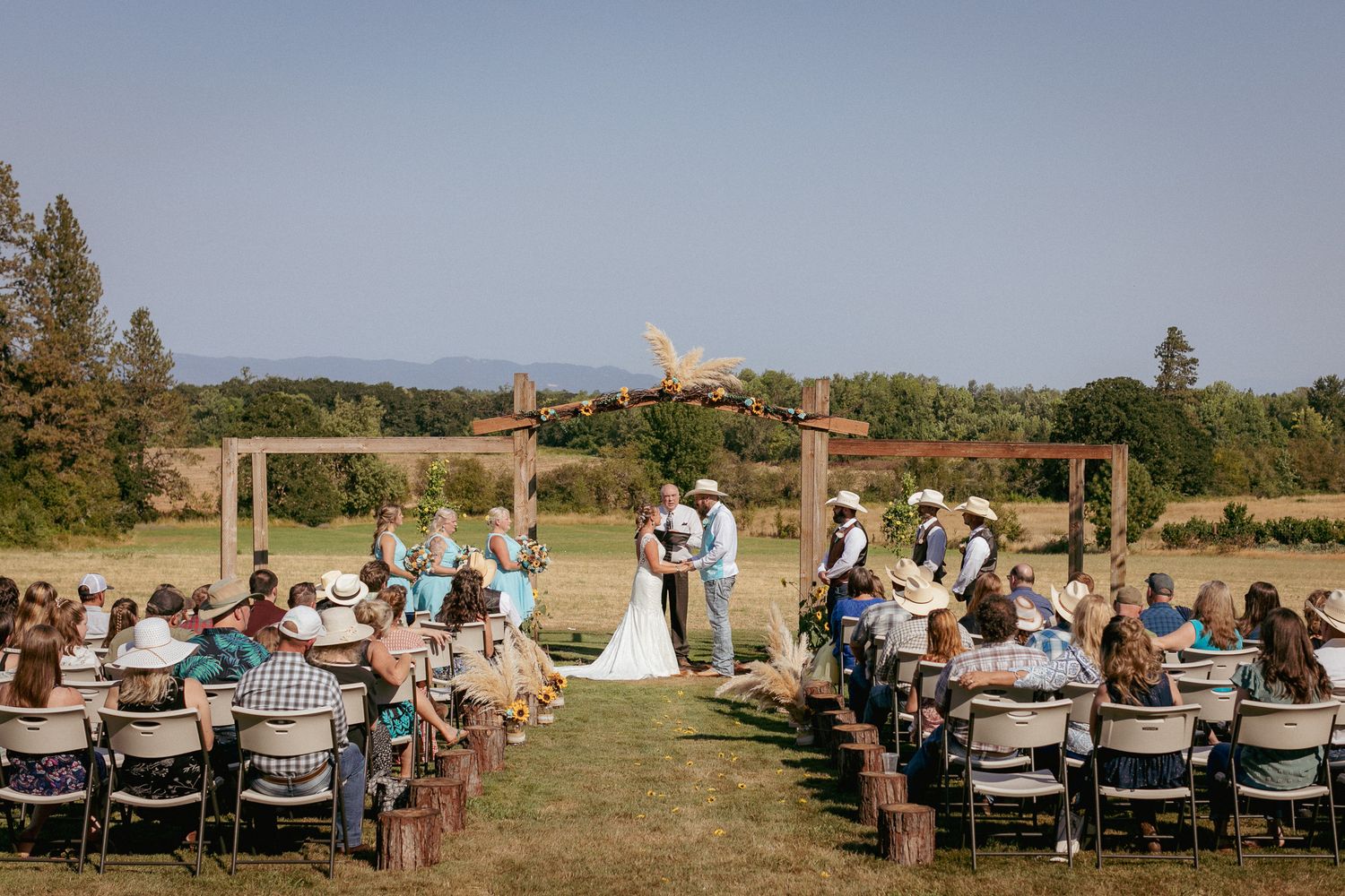 Guests seated outdoors at vineyard wedding ceremony with wooden arch and mountain view.