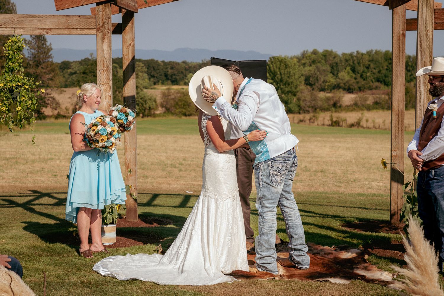 Outdoor Oregon country chic wedding first kiss, groom playfully covering bride’s face with his cowboy hat after the I dos. Romantic moment.