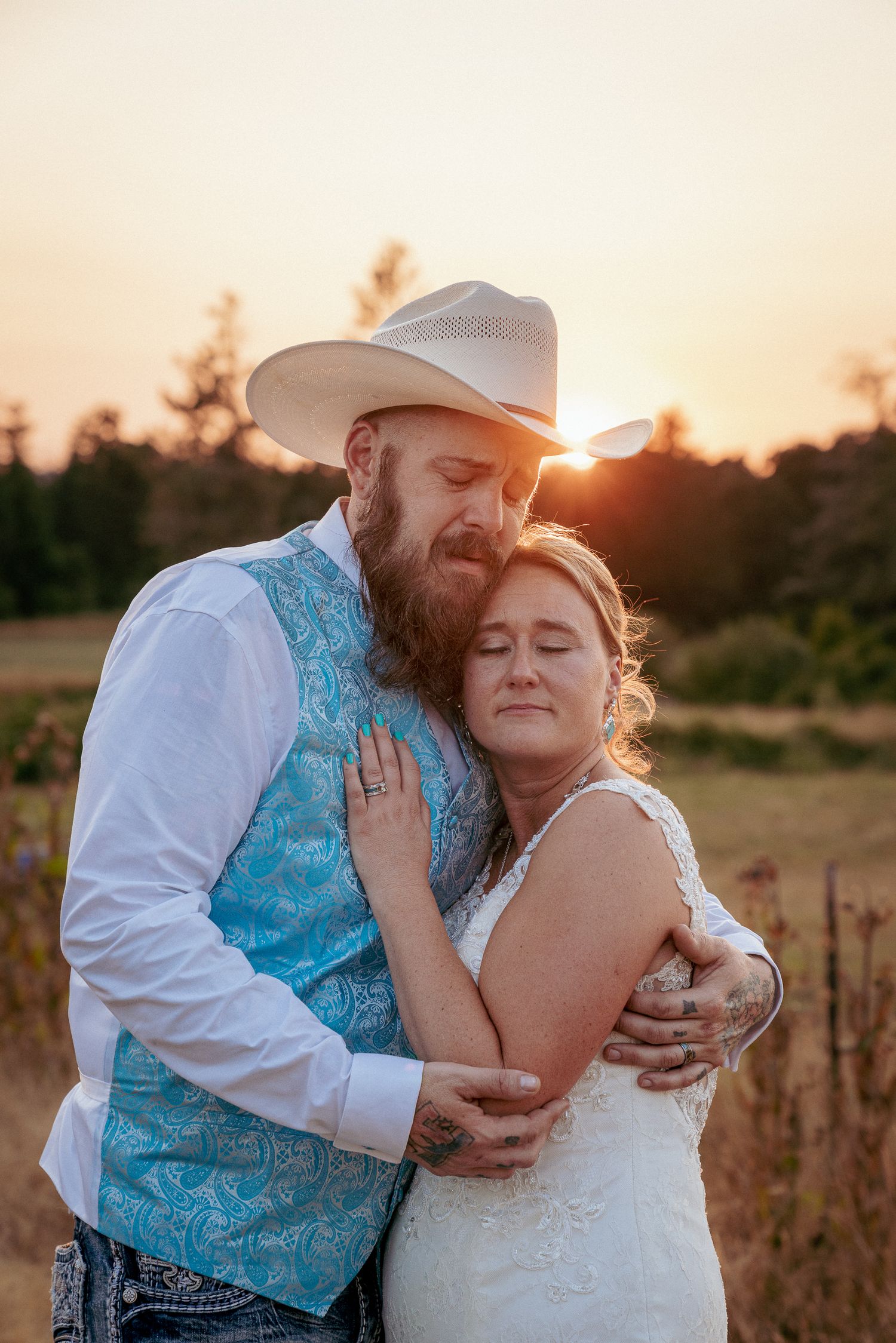 Groom in cowboy hat hugs bride at golden hour in country wedding setting.