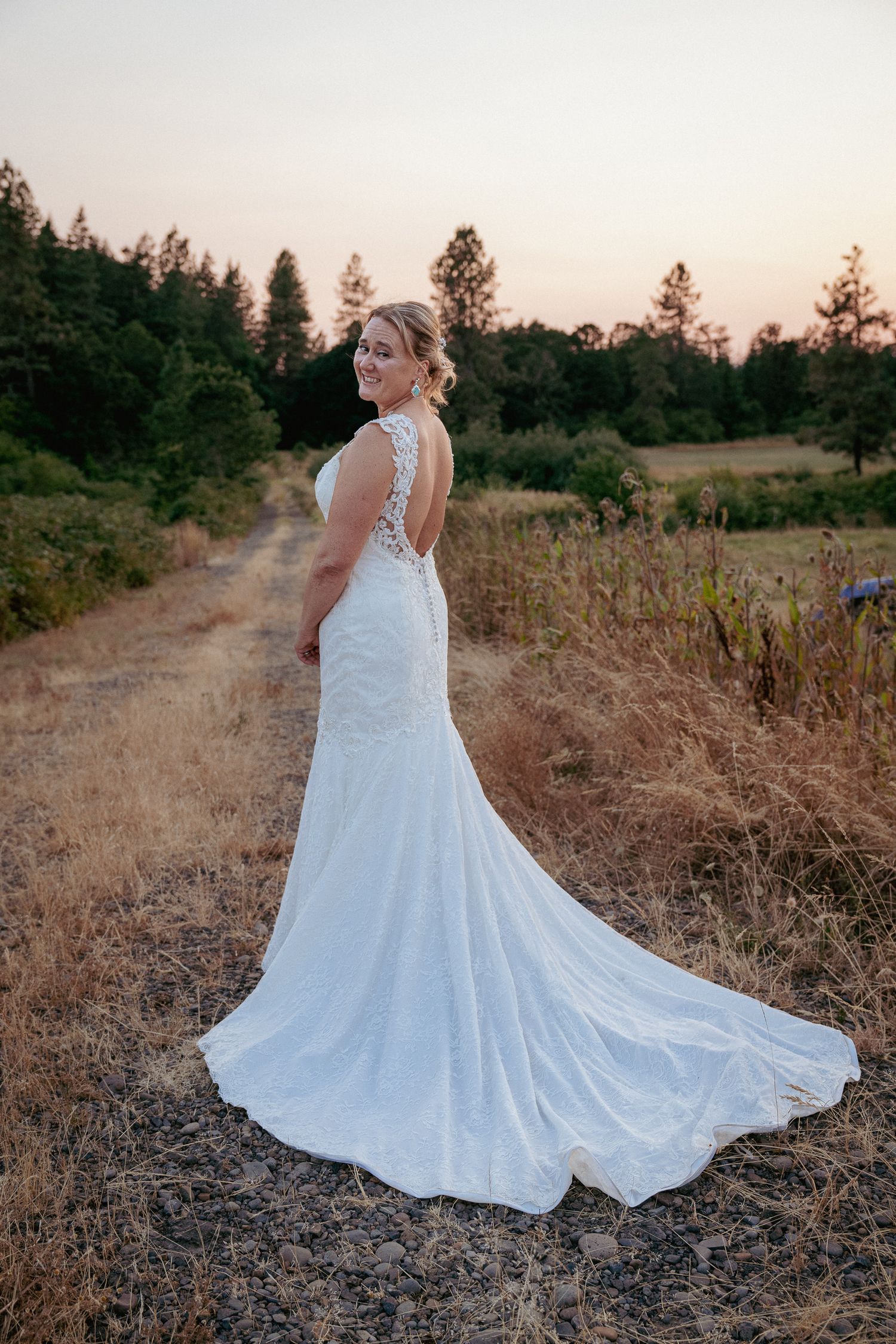 Bride walking alone down rustic path at sunset in fitted lace gown.