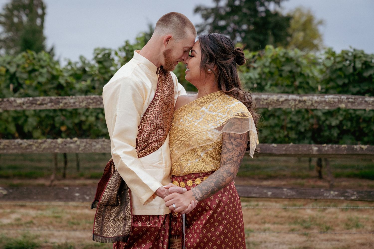 Interracial couple in traditional Southeast Asian wedding attire gazing into each other's eyes, holding hands outdoors with greenery in the
