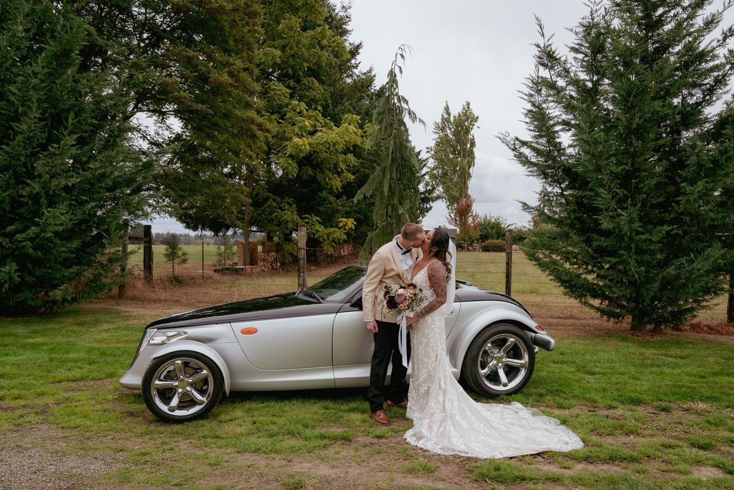 Bride and groom kiss next to silver convertible car. Fun wedding photo with luxury vehicle.