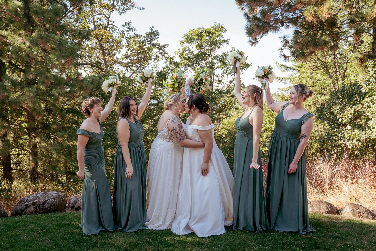Brides are kissing, bridesmaids in sage green dresses laughing together and cheering for the brides in the forest.