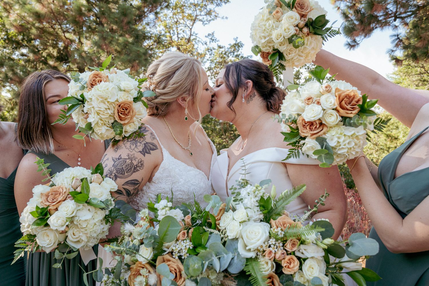 Two brides kiss, surrounded by bridesmaids holding bouquets in a wreath shape. LGBTQ+ wedding celebration with colorful flowers.