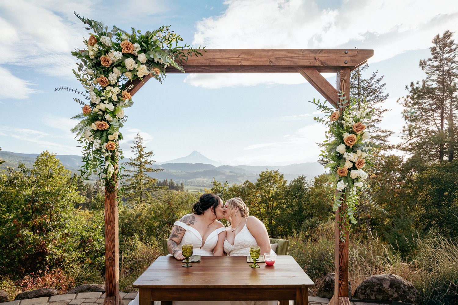Interracial lesbian couple kiss under floral arch, Columbia Gorge. With Mt Hood in the background