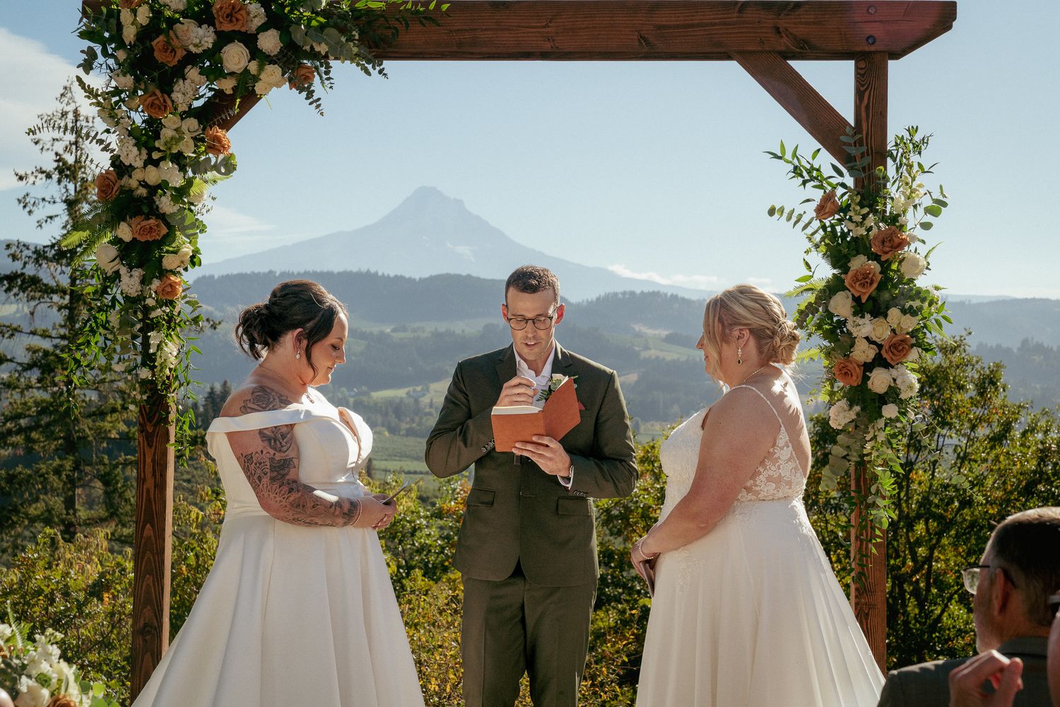 Two brides exchange vows with officiant, Mount Hood towering in the background. Intimate ceremony in Oregon's Cascade Mountains.