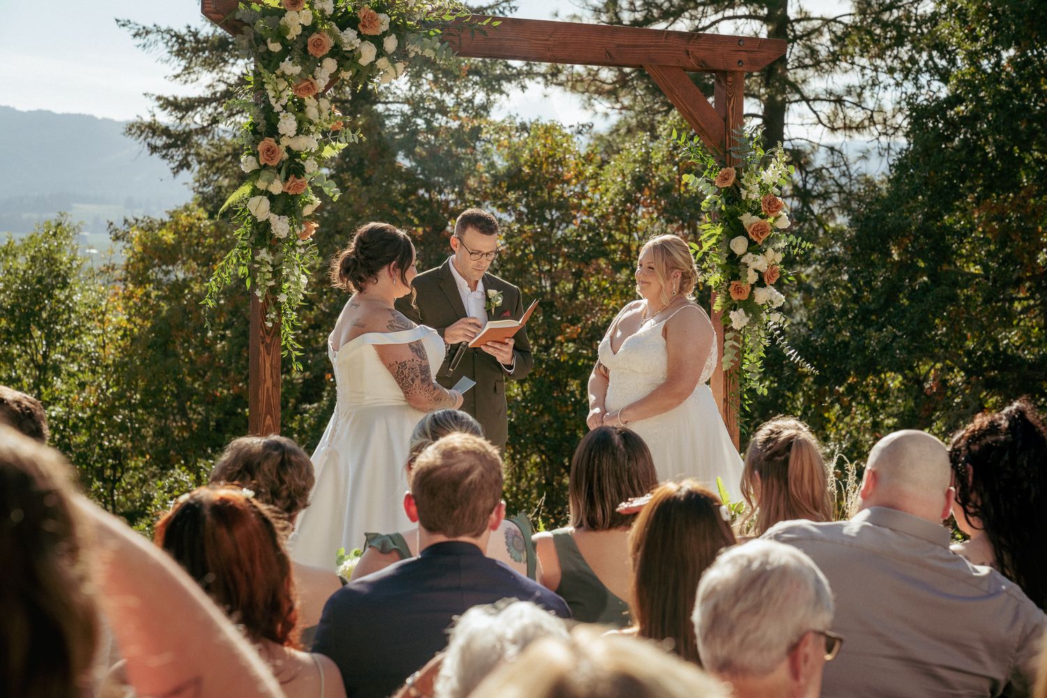 The brides exchange vows at an outdoor wedding ceremony under floral arch with guests watching.