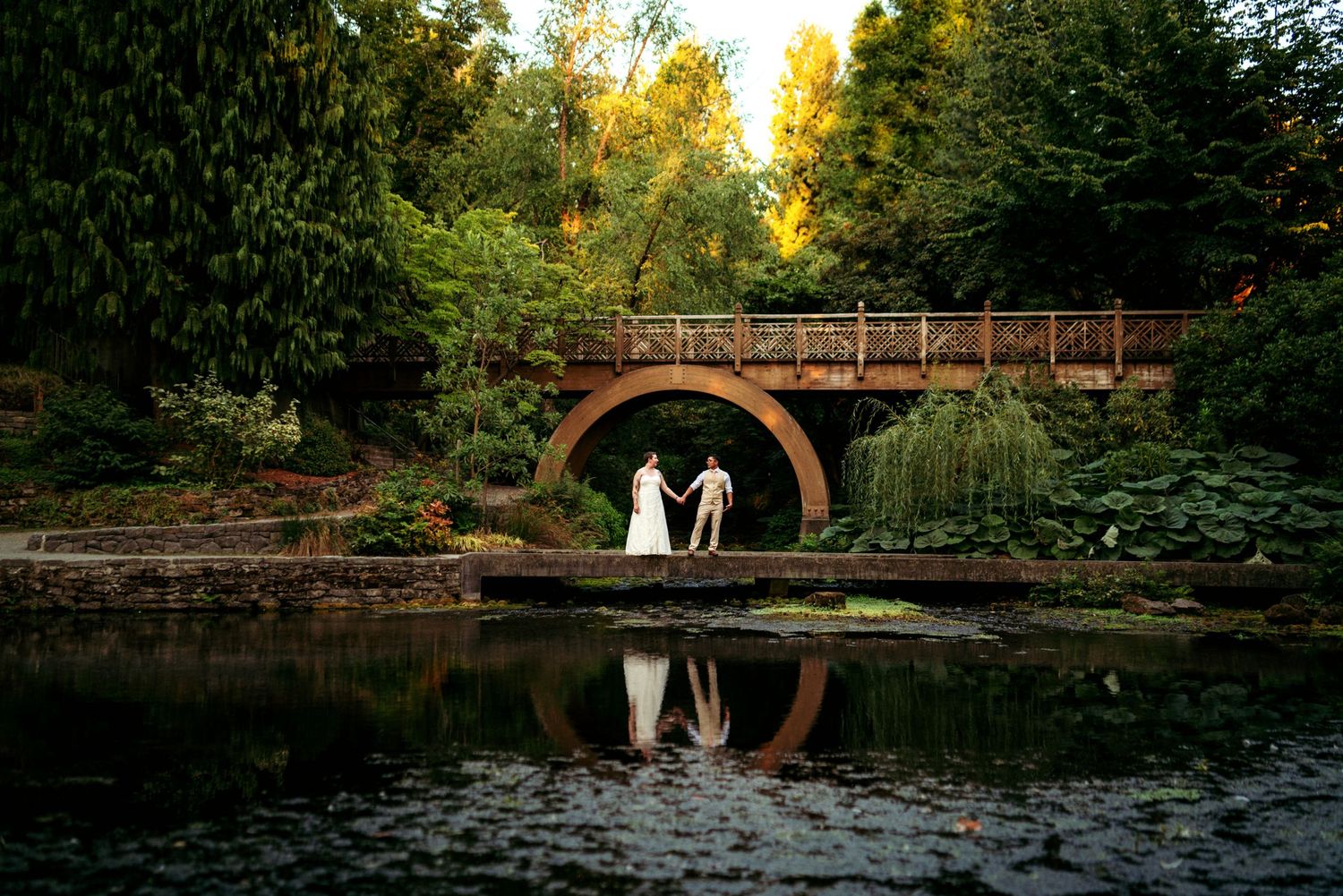 Bride and Groom holding hands on a picturesque arched bridge over a lily pond, surrounded by lush greenery and golden evening light.