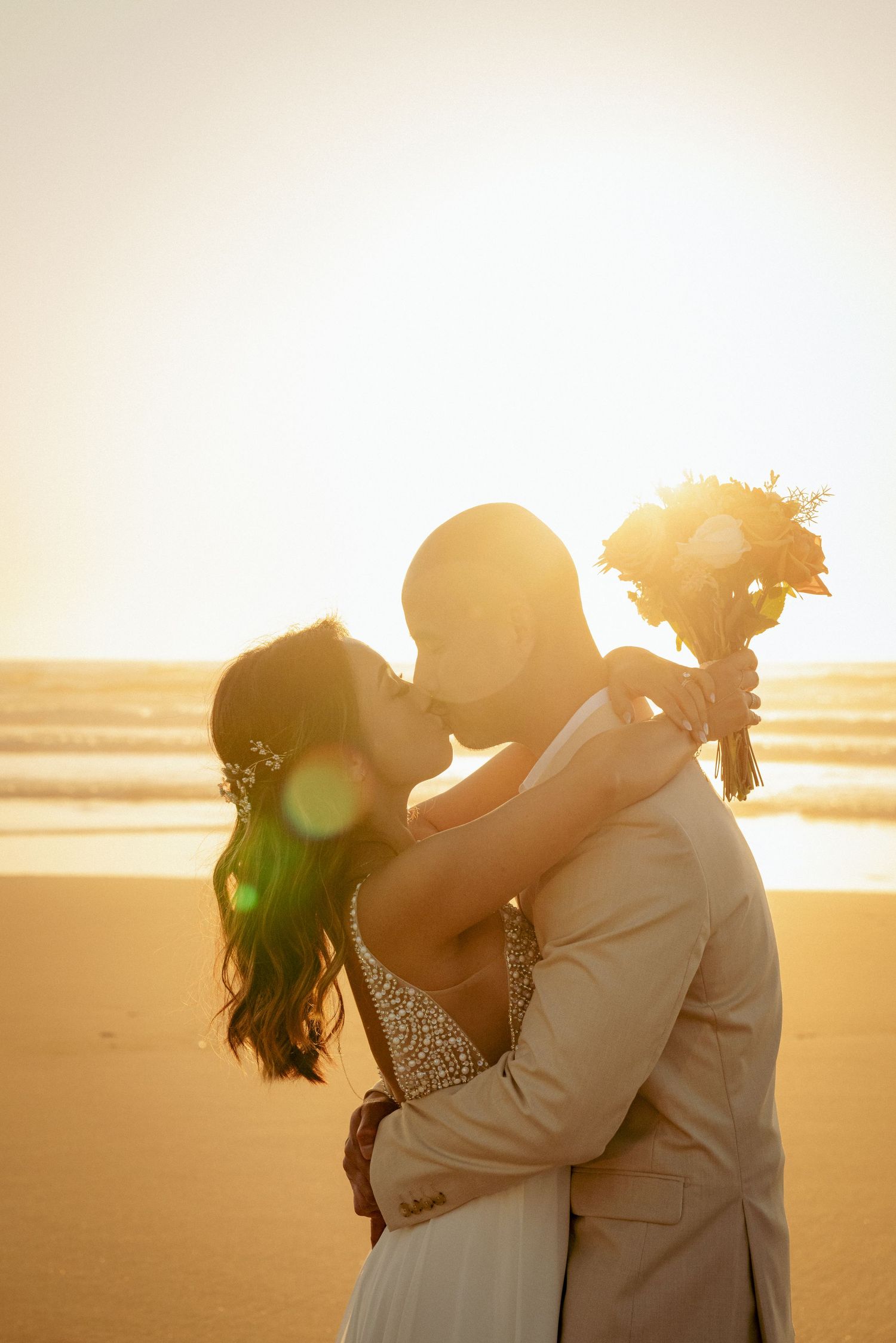 Couple embracing and kissing with sunset behind the on the beach, with the bride holding a bouquet and the golden light glowing around them.