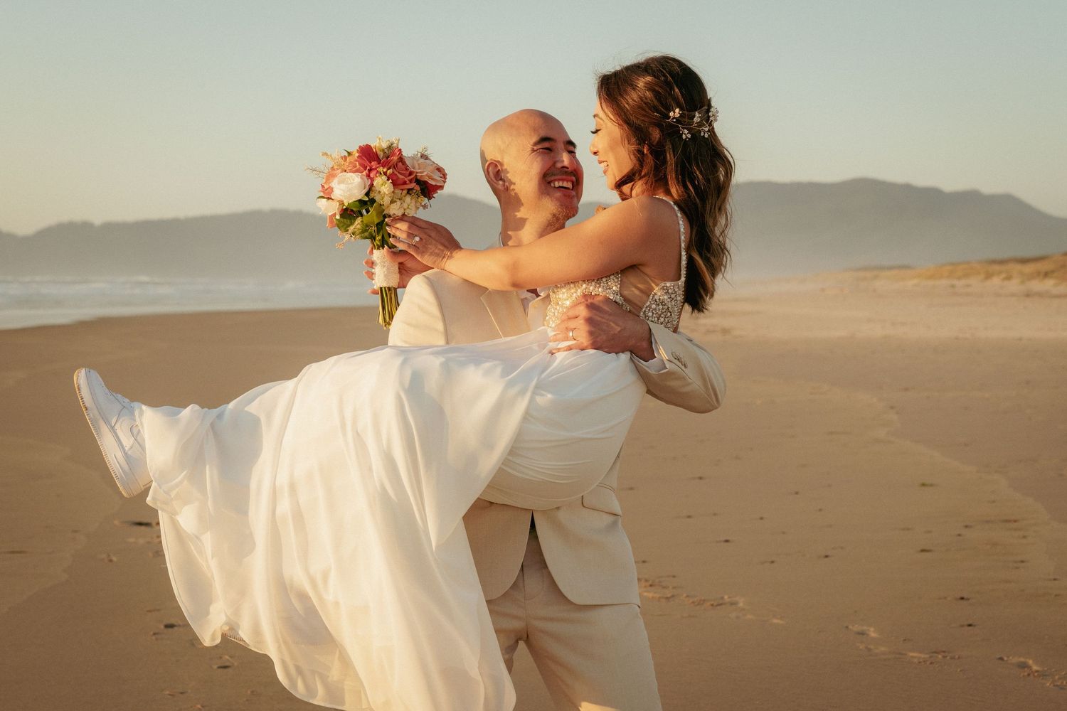 Groom lifts bride on sandy beach while she holds bouquet. Joyful post-ceremony moment.