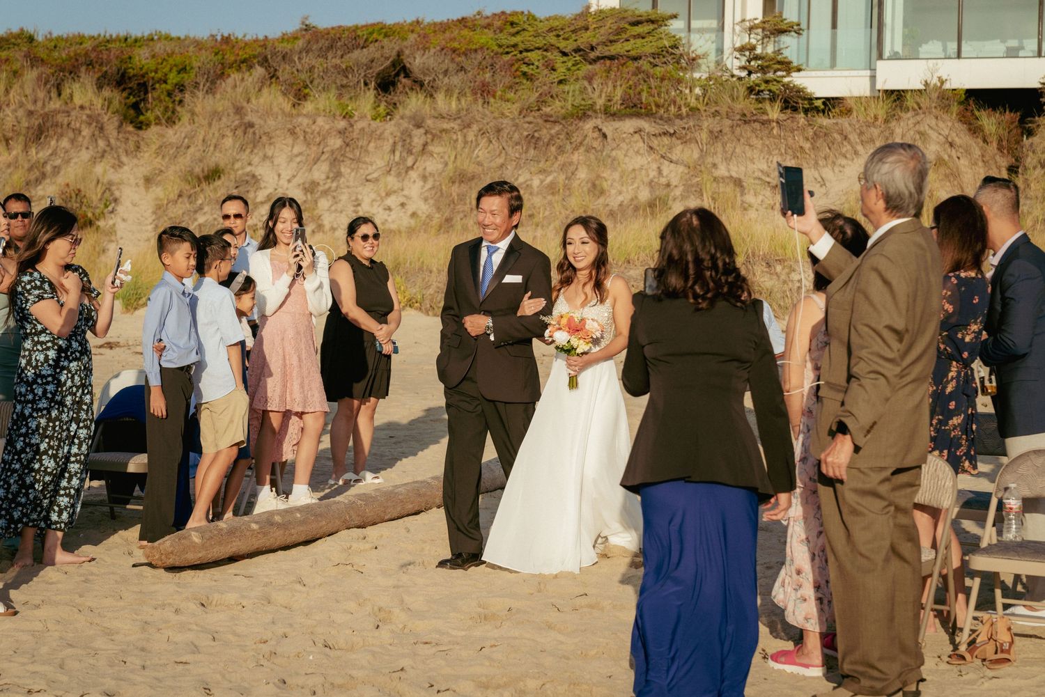 Bride walks down the aisle with her father at Oregon Coast wedding, guests standing. Asian family beach ceremony.