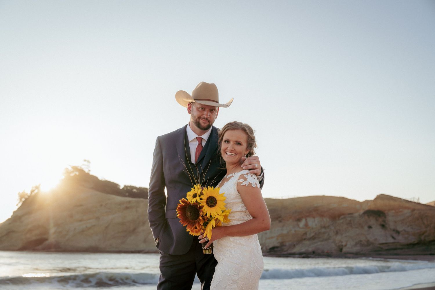 Bride and groom with sunflowers pose by Oregon ocean cliffs, with the groom in a cowboy hat and sunset light behind them.