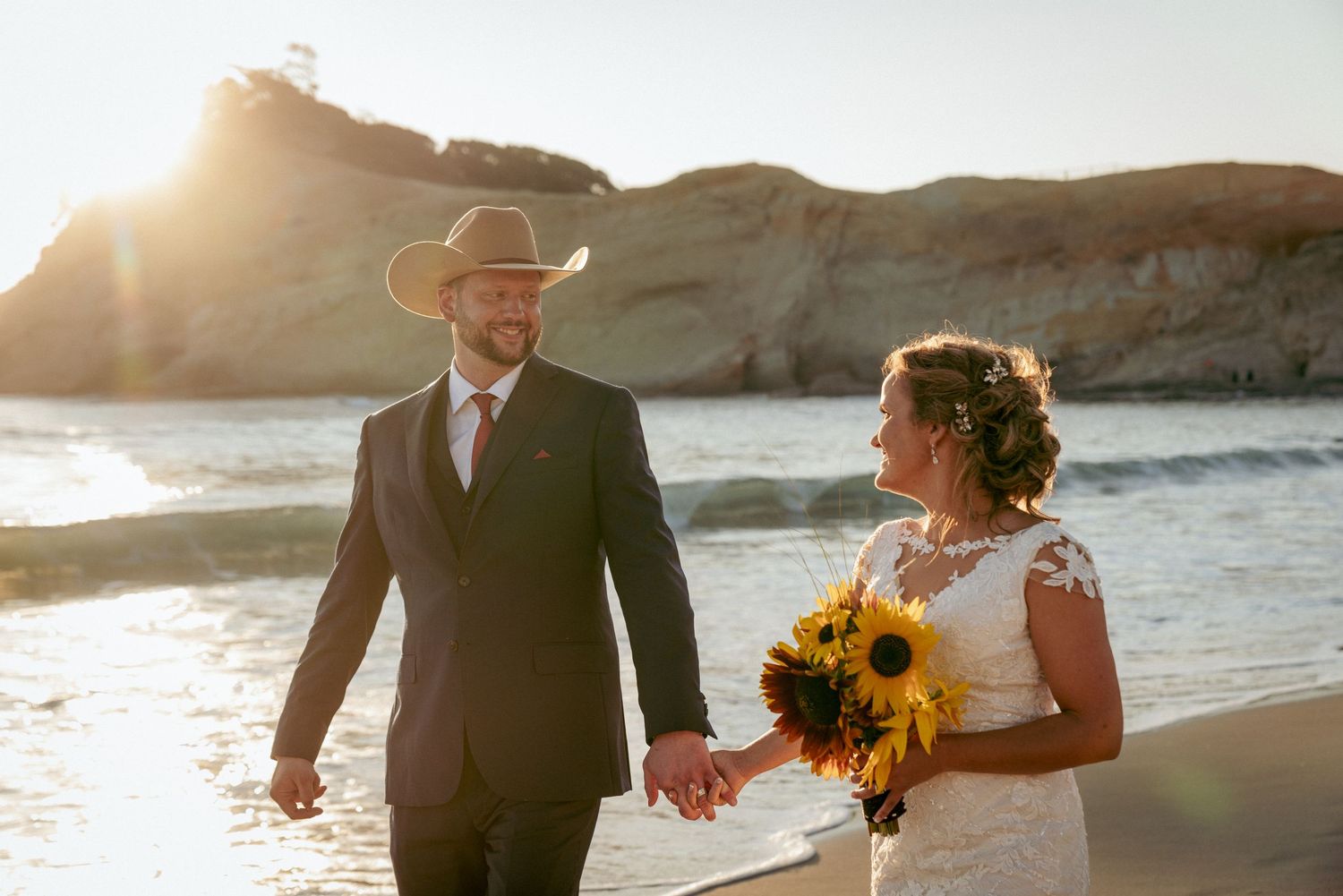 Bride and groom walk along Oregon Coast beach at sunset, with sunflower bouquet and groom in cowboy hat. Cliffs in the background.
