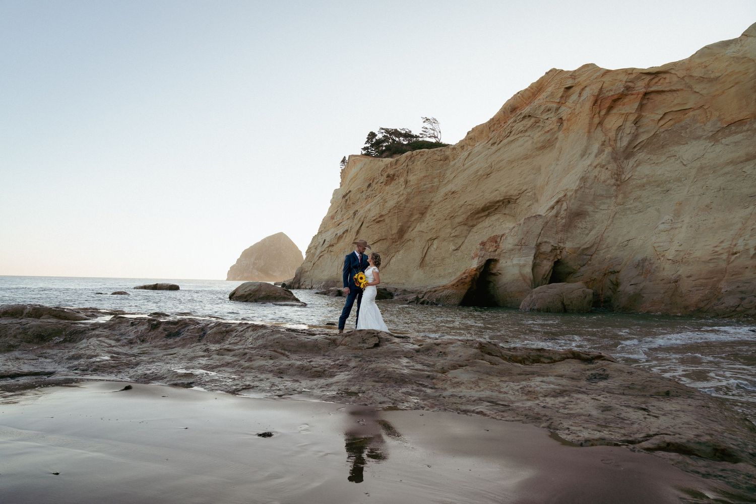 Groom and bride standing on a huge rock in ocean at Oregon Coast beach with rocky cliffs in the background.