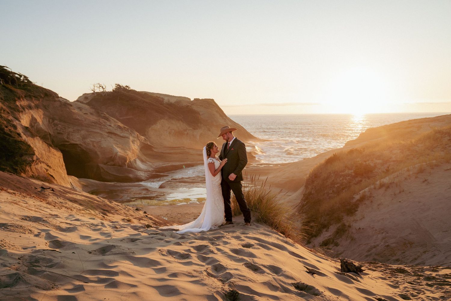 Bride and groom atop sand hill with Pacific Ocean, cliffs, and sunset behind them. Stunning wide scenic coastal wedding moment.