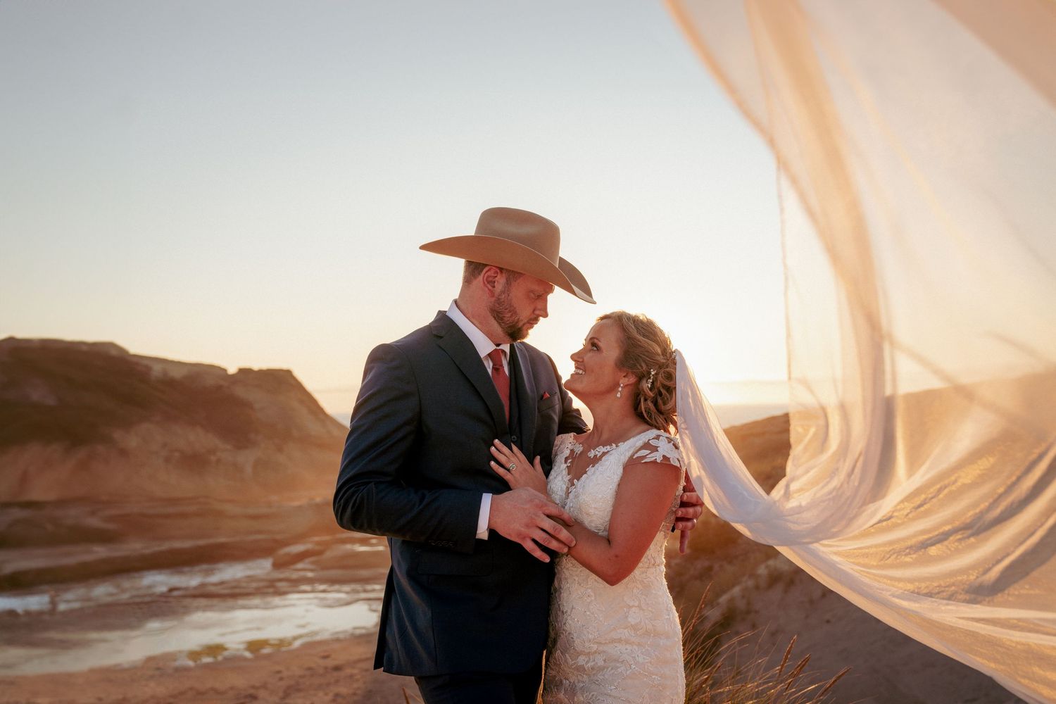 Close-up of bride and groom gazing at each other; groom wears a cowboy hat, bride’s veil flows toward the viewer. Intimate wedding moment.