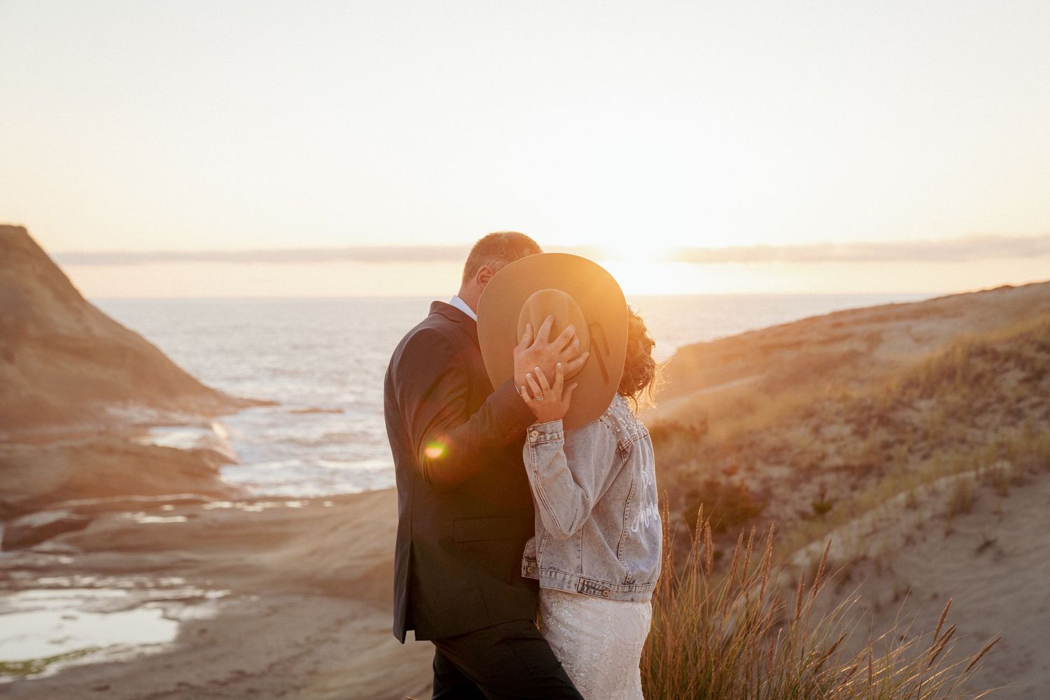 Bride and groom kissing, but covering their his with cowboy hat, at Oregon Coast dunes, golden sunset romance.