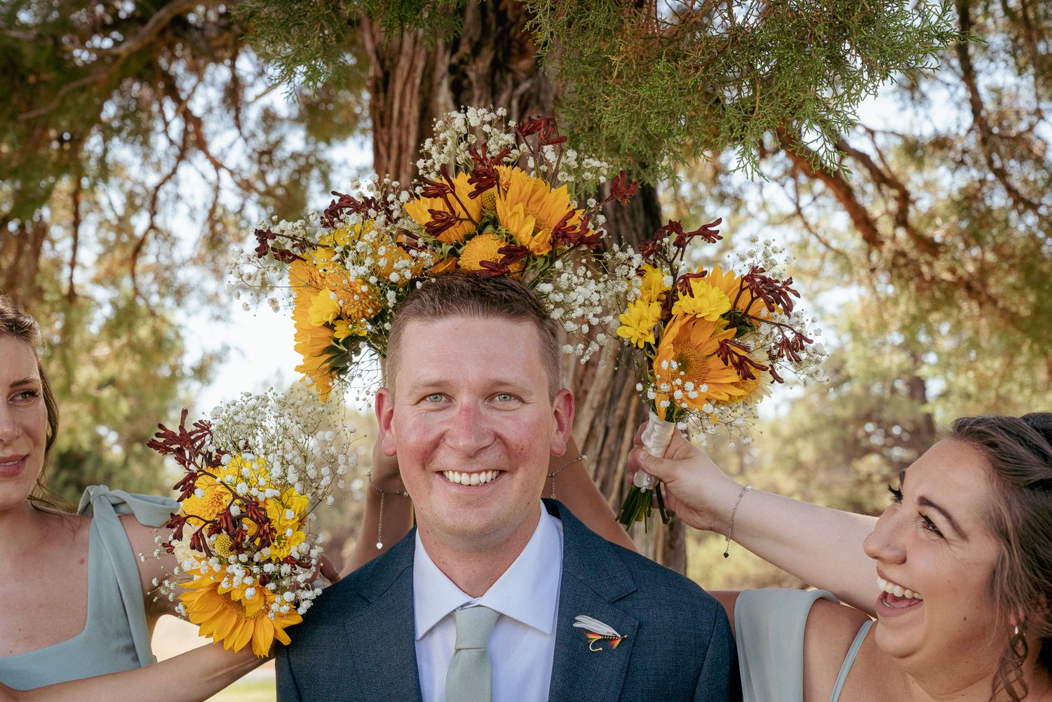 Groom surrounded by bridesmaids and their bouquets of sunflower creating a wreath around him, smiling before ceremony. Rustic summer wedding