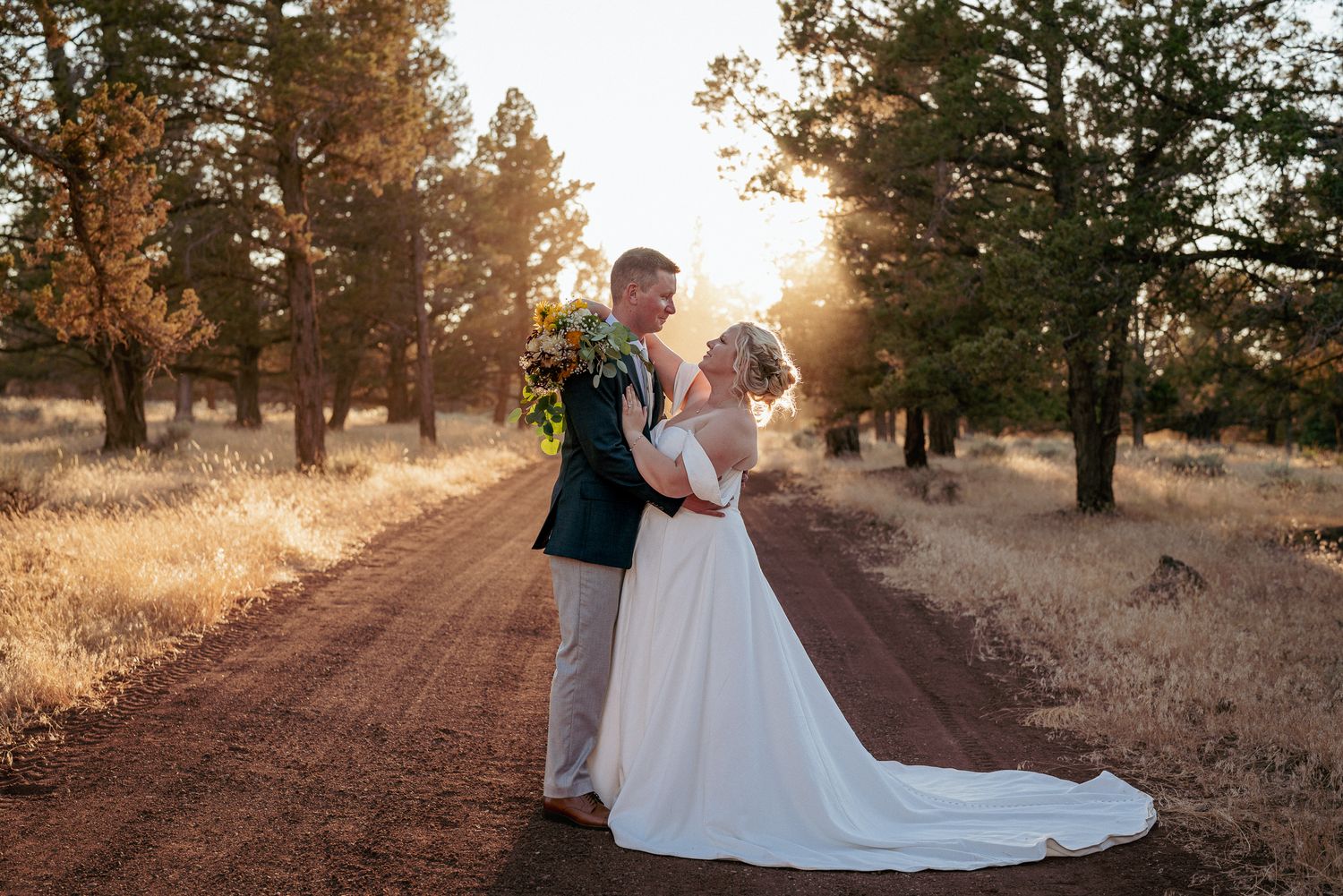 Bride and groom gaze into each other’s eyes on a country road at sunset, surrounded by dry grass and trees. Romantic outdoor moment.