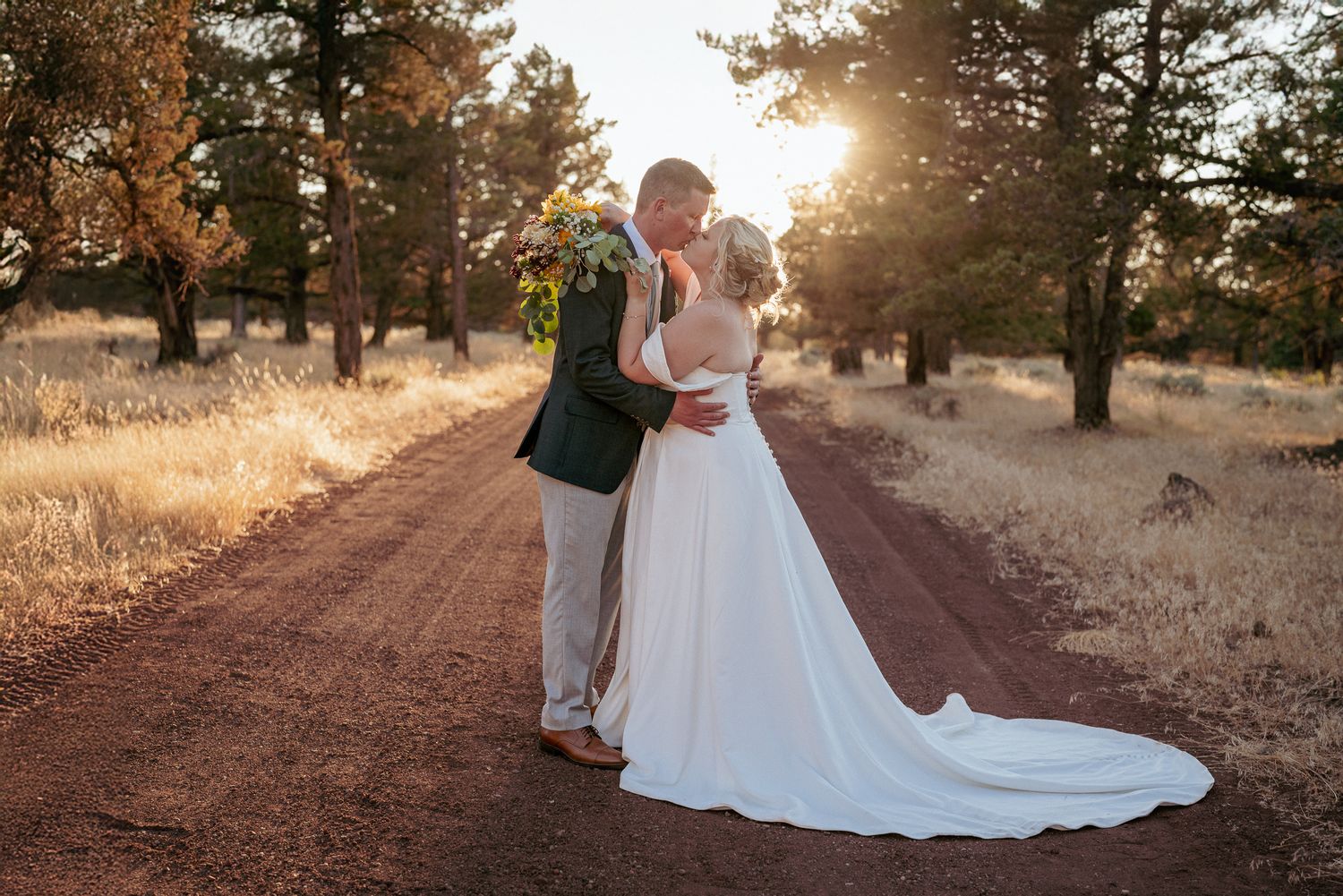 Bride and groom kiss on a forest road in Central Oregon with golden sunset light glowing behind them during their romantic outdoor wedding.
