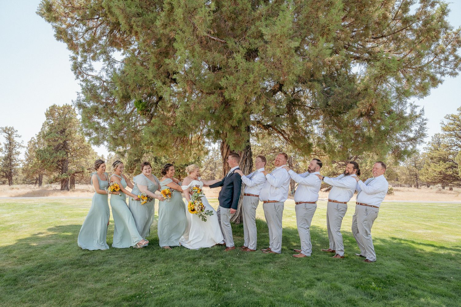 Bridal party playing tag of war on lush green lawn. Fun group photo with colorful bridesmaid dresses. Central Oregon Wedding