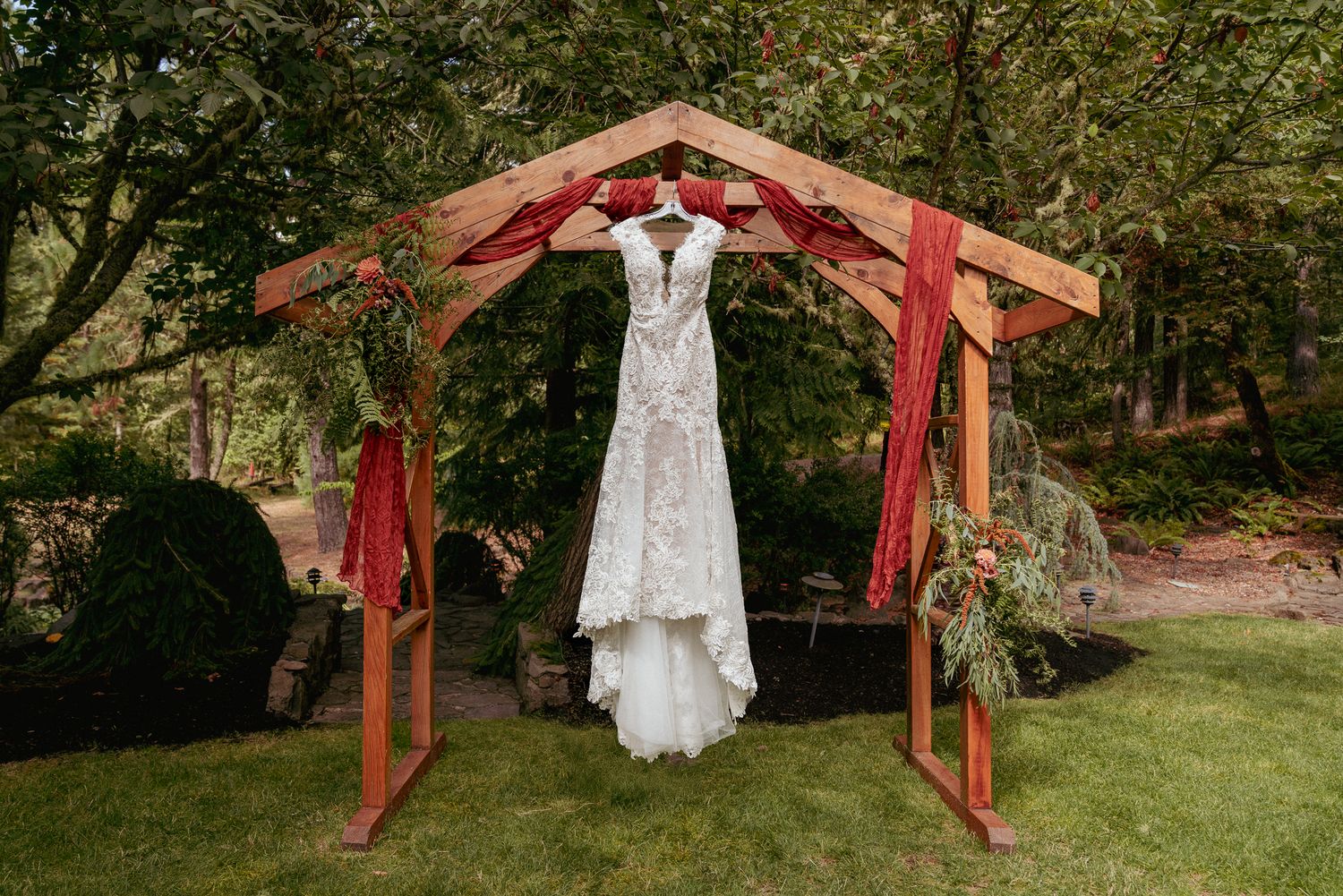Lace wedding gown hanging under a wooden arbor decorated with greenery and rust-colored drapery.