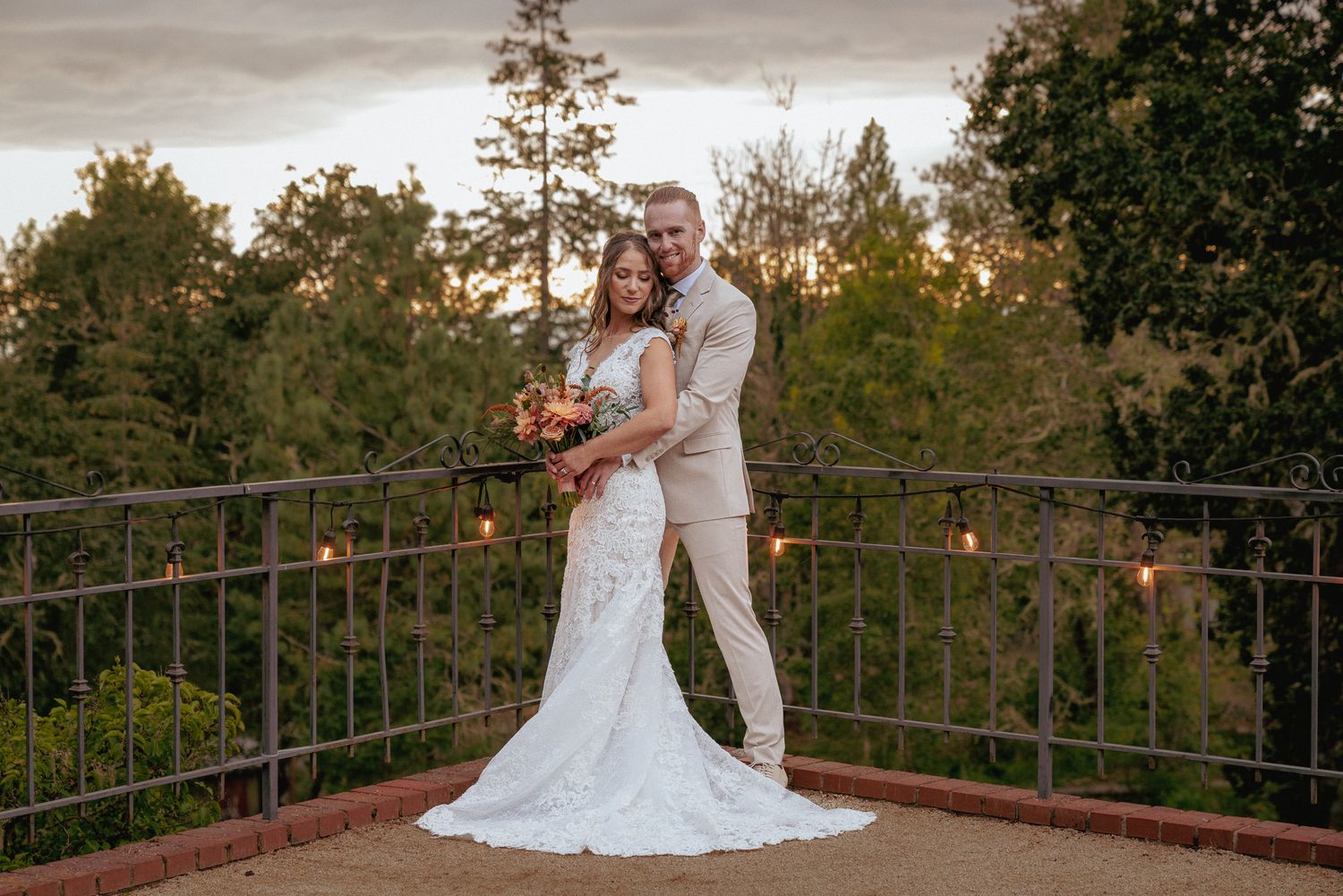 Groom hugs bride from behind on patio overlooking trees at sunset. Warm, intimate moment during outdoor wedding.