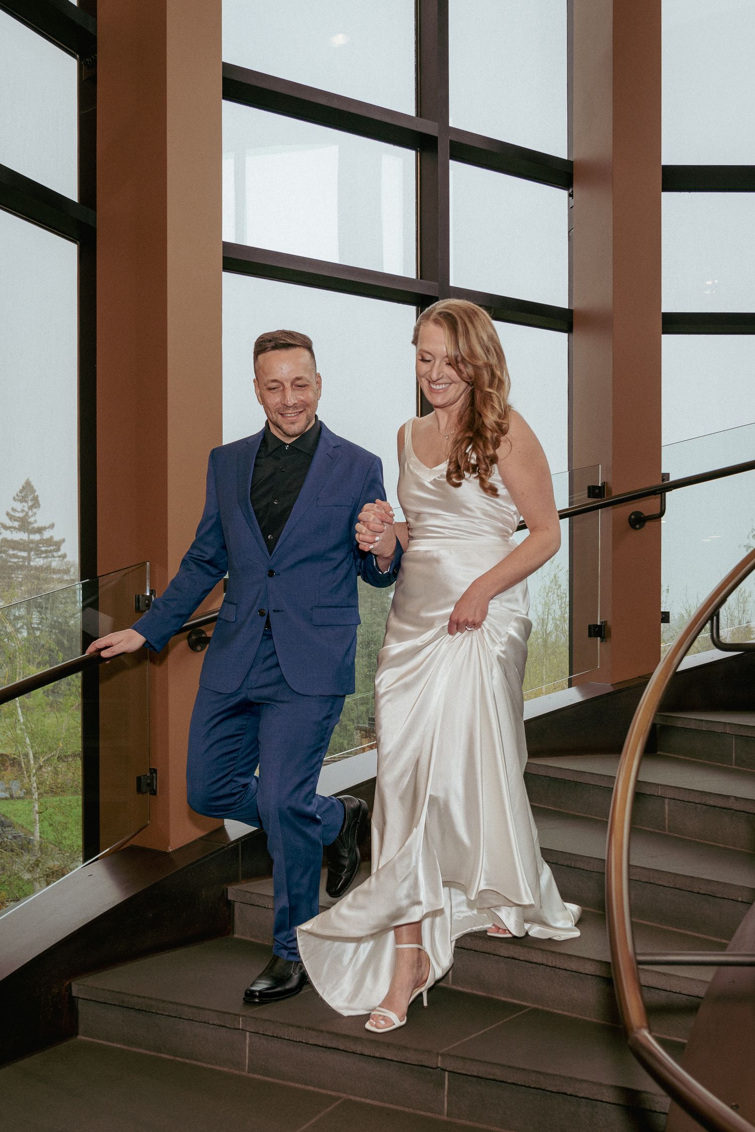 Bride and groom smile and walk down a modern indoor staircase during an elegant Portland Metro Area wedding.