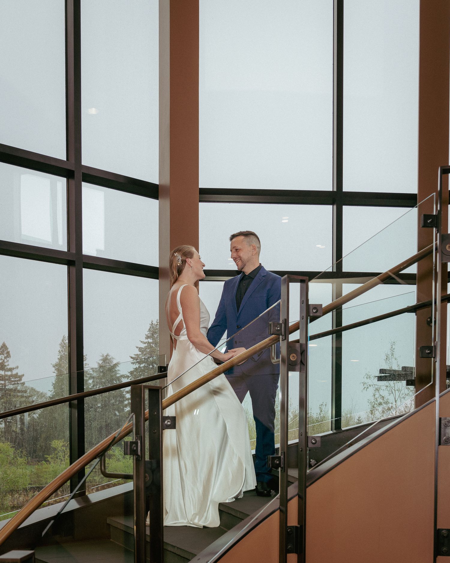 Bride and groom hold hands on a modern staircase with tall glass windows during a stylish Pacific Northwest wedding.