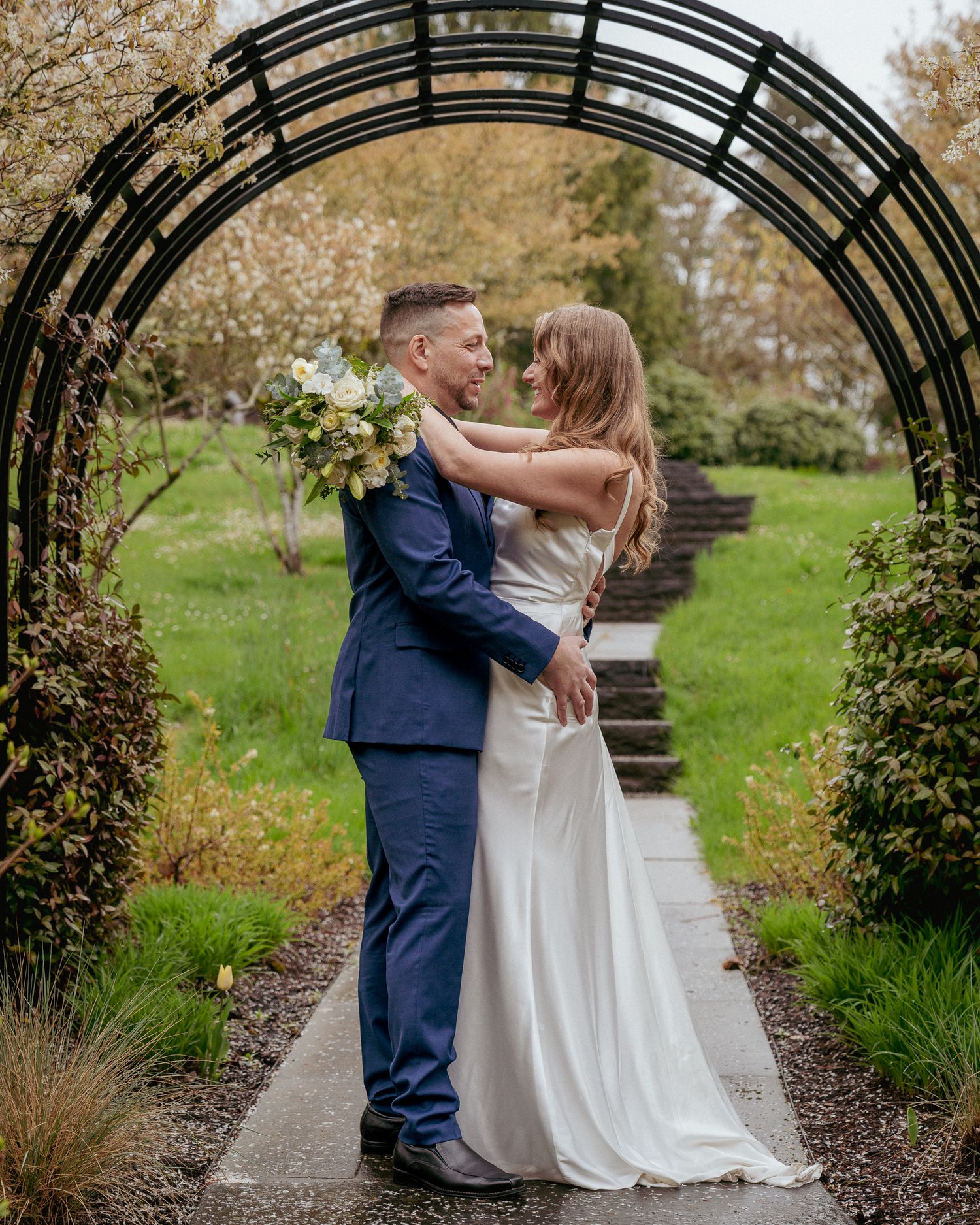Bride and groom hug and gaze into each other’s eyes under a metal arch, surrounded by lush greenery. Intimate wedding moment outdoors.