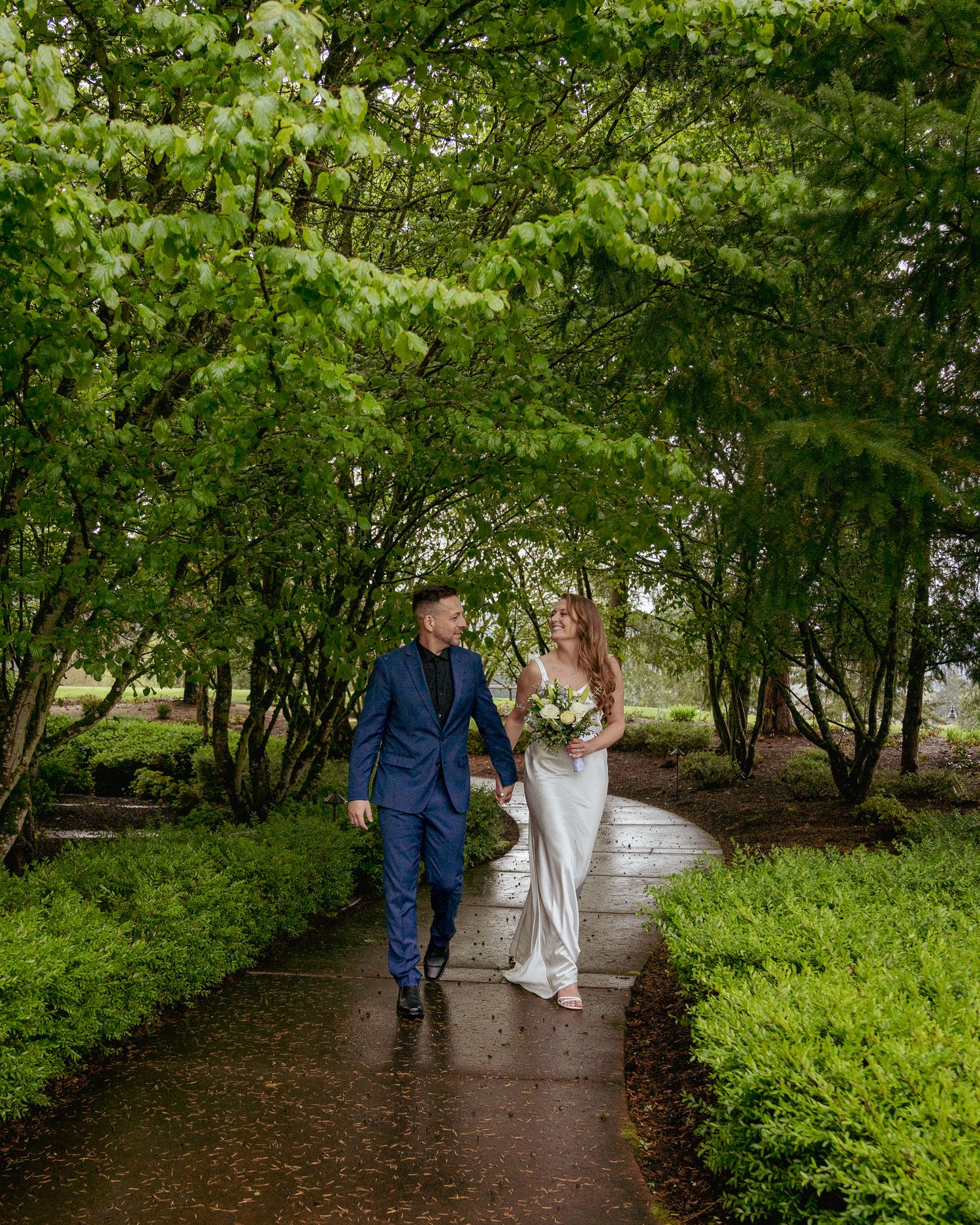 Bride and groom walk toward the camera on a paved path surrounded by trees and greenery, smiling and looking at each other.