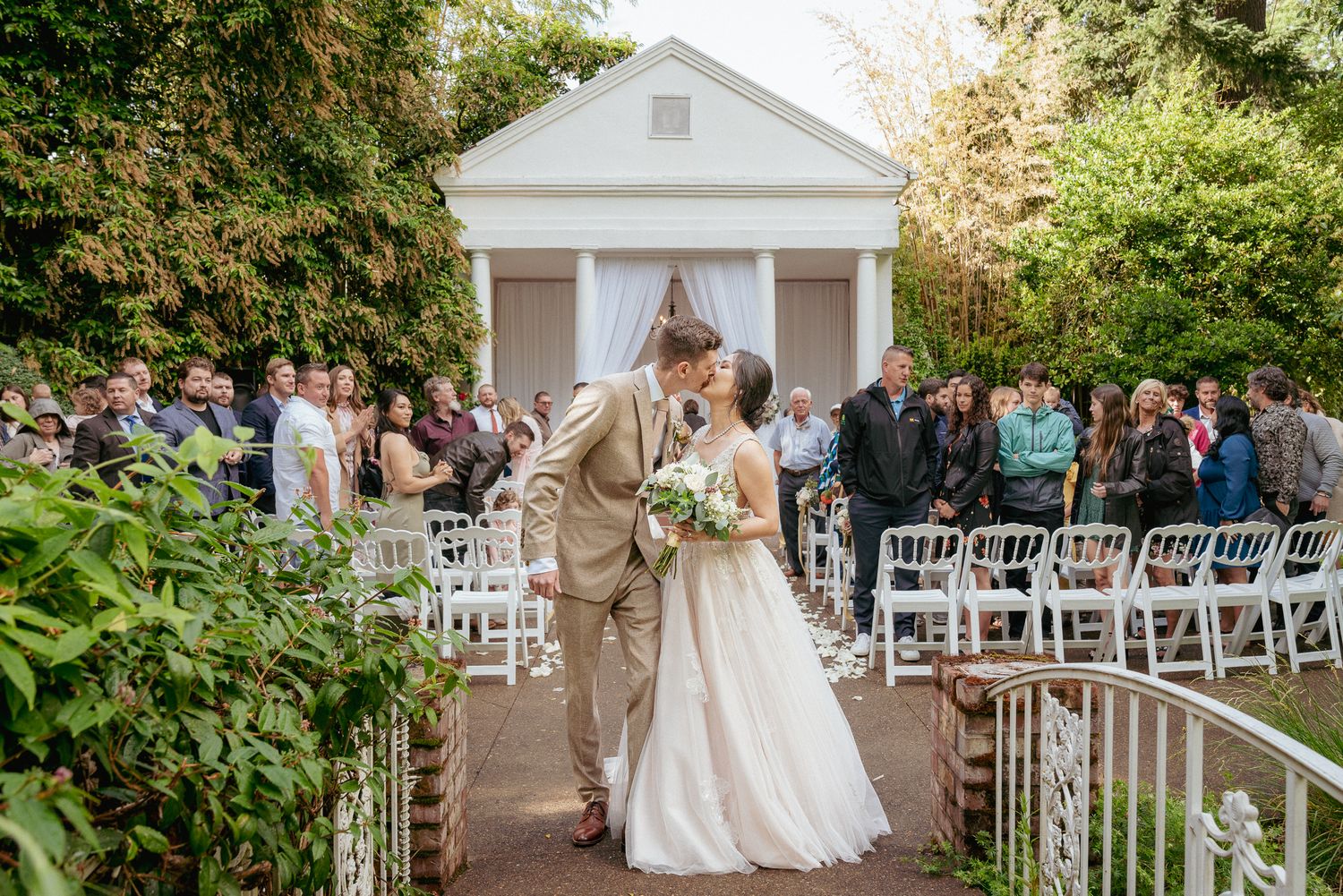Bride and groom share a kiss while walking down the aisle after their outdoor ceremony at a garden venue.