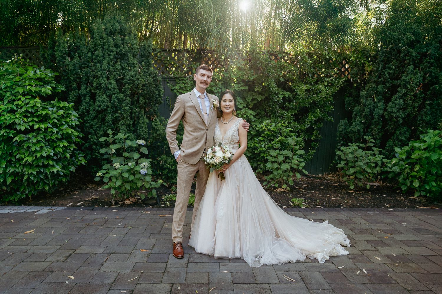 Bride and groom stand beneath tall trees in golden light. Romantic forest wedding portrait.