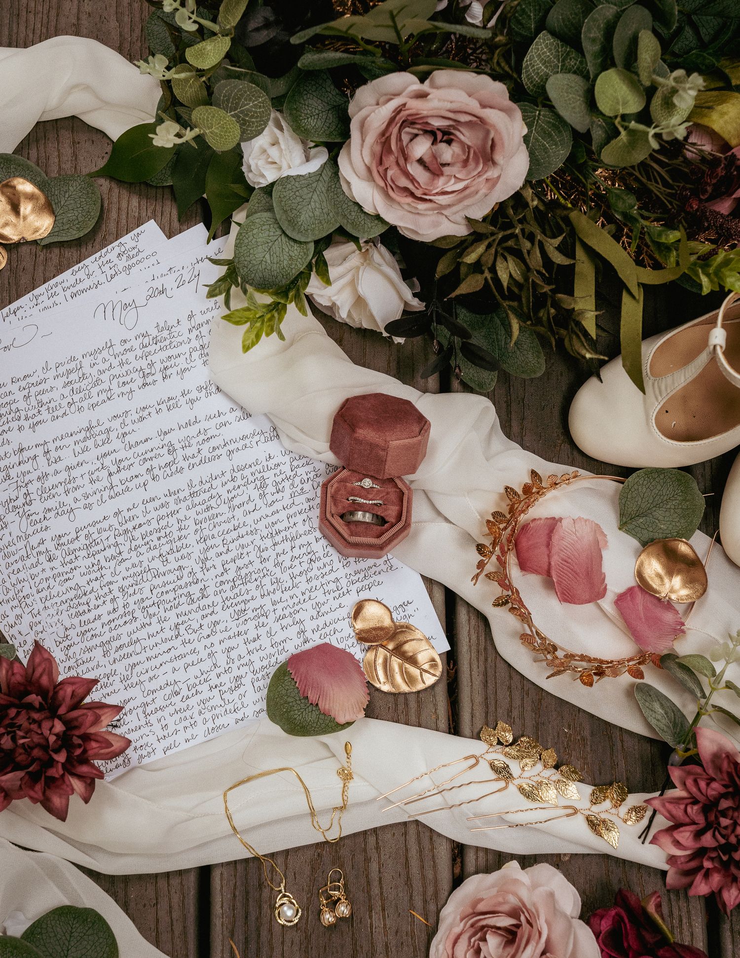 Flat lay of romantic wedding details including handwritten vows, velvet ring boxes, blush roses, and bridal accessories on soft fabric.