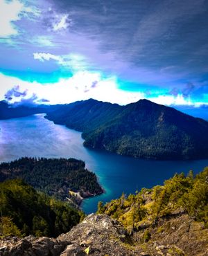 Aerial view of pristine mountain lake surrounded by forested peaks under dramatic sky.