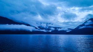 Moody blue landscape with dramatic storm clouds over mountain lake.