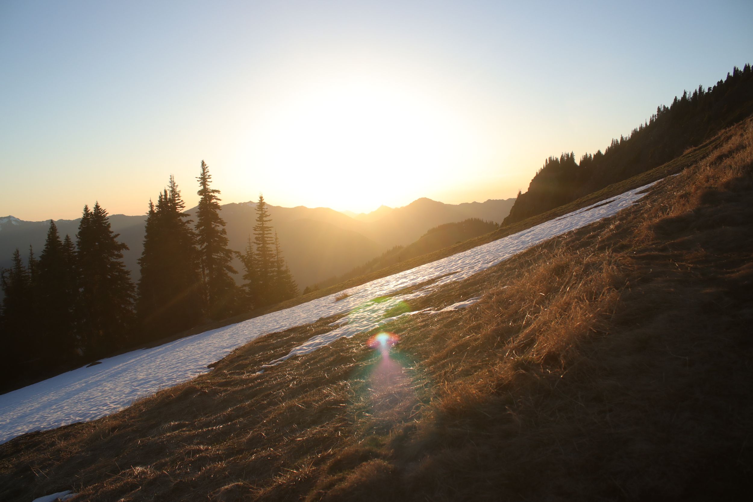 Scenic mountain landscape at sunset with snow patches and evergreen trees.